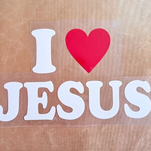 Könnte beinhalten: Weiße Vinylbuchstaben, die "Ich liebe Jesus" buchstabieren, mit einem roten Herzen zwischen den Wörtern "Ich" und "liebe".