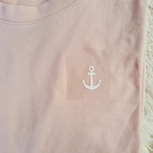 Könnte beinhalten: Hellrosa T-Shirt mit weißem Anker-Logo auf der Brust. Das Shirt hat einen Rundhalsausschnitt und ist aus weichem Material. Das Anker-Logo ist ein einfaches, nautisches Design.