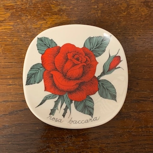 May include: A white ceramic dish featuring a red rose and green leaves. The dish is inscribed with the words "rosa baccara" at the bottom.