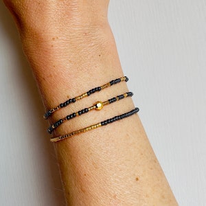 Op de afbeelding: Drie gelaagde armbanden met zwarte en gouden kralen. De armbanden zijn gestapeld op de pols van een persoon. De bovenste armband heeft een gouden kraal in het midden.