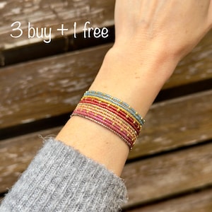 Può includere: Braccialetto di perline con più fili nei toni del rosa, rosso, oro e blu. Il testo "3 buy + 1 free" è visibile nell'immagine. Il braccialetto è indossato su un polso, con una manica di maglione grigio in primo piano.