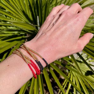 Op de afbeelding: Een pols versierd met vier armbanden van kralen. De armbanden hebben gouden, rode en zwarte kralen. De gouden kralen zijn het meest prominent, met de rode en zwarte kralen die een vleugje kleur toevoegen. De armbanden worden gedragen op een pols tegen een achtergrond van groen gebladerte.