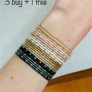 Peut inclure: Plusieurs bracelets de perles de différentes couleurs, dont noir, or, blanc et rose. Les bracelets sont ornés de petites perles dorées. Le texte de l'image indique "3 buy + 1 free".