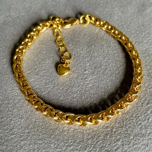 Op de afbeelding: Een gouden kettingarmband met een hartvormige hanger. De ketting is in een gevlochten ontwerp.