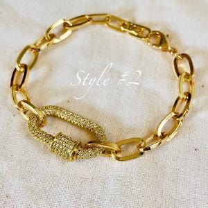 Puede incluir: Pulsera dorada con un cierre grande y adornado y eslabones de cadena ovalados. El cierre está incrustado con pequeñas piedras brillantes. El texto "Style #2" está escrito en cursiva en el fondo.