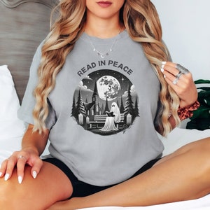 Op de afbeelding: Grijs t-shirt met de tekst "READ IN PEACE" boven een zwart-witte afbeelding. De afbeelding toont een geest die een boek leest op een bank, met een kerk en volle maan op de achtergrond.