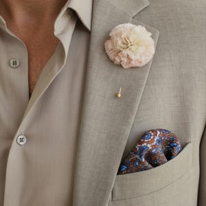 May include: A light beige blazer with a matching button-down shirt. A light pink flower lapel pin and a gold pin are attached to the blazer. A patterned pocket square with blue and brown accents is in the pocket.