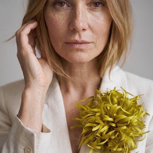 Puede incluir: Un broche floral grande y redondo de color chartreuse que presenta numerosos pétalos finos y rizados en forma de cinta. El broche está prendido en la solapa de una americana de color beige claro con textura y tres botones visibles en el puño.