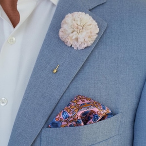 Könnte beinhalten: Hellblauer Blazer mit weißem Hemd darunter. Eine cremefarbene Blumen-Reversnadel und eine goldene Krawattennadel sind am Blazer befestigt. Ein Einstecktuch mit rosa und blauem Muster befindet sich in der Brusttasche.
