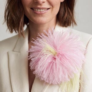 Könnte beinhalten: Eine große, flauschige rosa Federbrosche mit gelben Akzenten ist an einem cremefarbenen Blazer befestigt. Die Brosche ist der Blickfang und verleiht dem Outfit einen Farbtupfer und Textur. Die Gesamtästhetik ist elegant und stilvoll.
