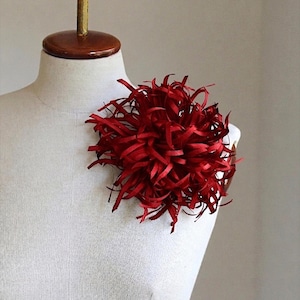 Puede incluir: Una vibrante flor de tela roja, posiblemente un broche o un adorno decorativo, está prendida a un maniquí blanco. La flor tiene un diseño texturizado con numerosos pétalos finos en forma de cinta que irradian hacia afuera.