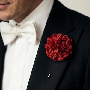Op de afbeelding: Een close-up van een formele outfit, met een zwarte smokingjas, een witte vlinderdas en een opvallende rode bloem in de knoopsgat. Een gouden reversspeld is ook zichtbaar. De afbeelding straalt klassieke elegantie uit.