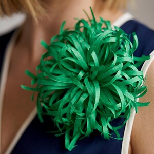 Puede incluir: Un vibrante accesorio floral verde, similar a una cinta. El accesorio tiene una forma grande y redonda con muchas cintas finas y rizadas que irradian hacia afuera. Está prendido a una prenda azul marino con ribete blanco. El efecto general es una pieza llamativa y audaz.