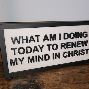 May include: A framed sign with the text "WHAT AM I DOING TODAY TO RENEW MY MIND IN CHRIST" in black letters on a white background. The frame is black, and the sign rests on a wooden surface.
