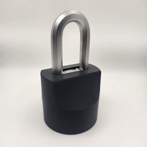 Puede incluir: Un candado negro con un grillete plateado en forma de U. El cuerpo del candado es cilíndrico y el grillete tiene un acabado plateado con una franja roja en el centro. El candado está sobre un fondo blanco.