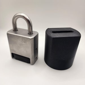 Puede incluir: Un candado plateado con una base de plástico negro. El candado tiene un grillete plateado y una tapa de plástico negro para la cerradura.