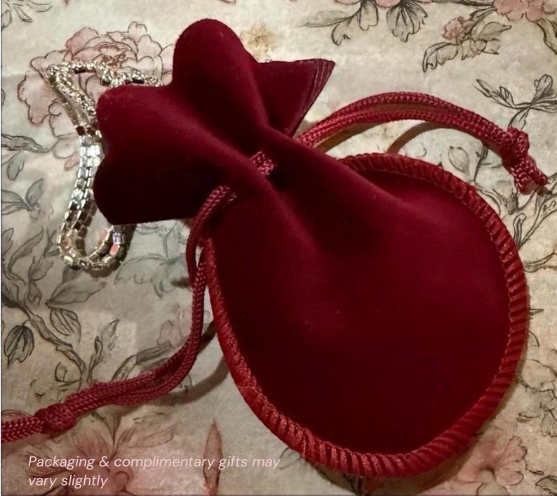 Pu&ograve; includere: Una collana d'argento &egrave; appoggiata vicino a un sacchetto di velluto rosso scuro con chiusura a coulisse. Il sacchetto &egrave; rifinito con un cordoncino rosso abbinato. Lo sfondo presenta un motivo floreale con colori tenui e smorzati. Il testo in basso dice "Packaging & complimentary gifts may vary slightly."
