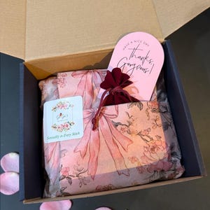Pu&ograve; includere: Una scatola di cartone aperta rivela una presentazione regalo. All'interno, carta velina con fiocchi rosa e motivi floreali avvolge il contenuto. Sono visibili anche un piccolo sacchetto bordeaux e un biglietto rosa con la scritta "Have a nice day, Thanks, Gorgeous!".