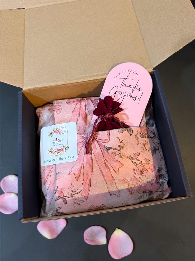 Pu&ograve; includere: Una scatola di cartone aperta rivela una presentazione regalo. All'interno, carta velina con fiocchi rosa e motivi floreali avvolge il contenuto. Sono visibili anche un piccolo sacchetto bordeaux e un biglietto rosa con la scritta "Have a nice day, Thanks, Gorgeous!".