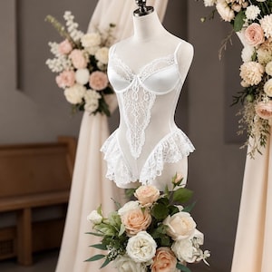 Pu&ograve; includere: Un body bianco in raso e pizzo esposto su un manichino. La lingerie presenta un delicato design in pizzo e finiture a balze. Il manichino &egrave; circondato da composizioni floreali con rose color pesca e bianche.