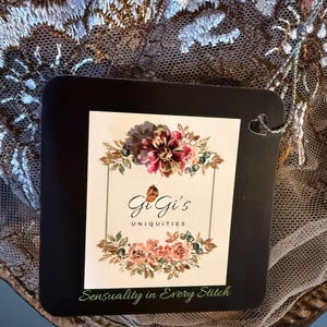Pu&ograve; includere: Una scheda quadrata nera con il testo "GiGi's Uniquities" e "Sensuality in Every Stitch" &egrave; mostrata. La scheda &egrave; decorata con un disegno floreale. La scheda &egrave; ambientata su uno sfondo di pizzo intricato, marrone e bianco.