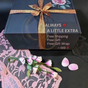 Pu&ograve; includere: Una scatola regalo grigio scuro con un nastro glitterato dorato e un design a rosa. La scatola ha il testo "ALWAYS A LITTLE EXTRA". Sono presenti anche petali di rosa rosa e una composizione floreale.