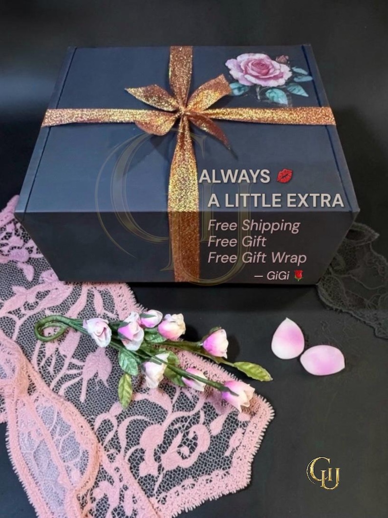Pu&ograve; includere: Una scatola regalo grigio scuro con un nastro glitterato dorato e un design a rosa. La scatola ha il testo "ALWAYS A LITTLE EXTRA". Sono presenti anche petali di rosa rosa e una composizione floreale.
