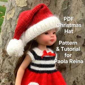 May include: A red and white knitted Santa hat with a white pom-pom on top. The hat is being worn by a doll with brown hair and a red and white striped dress. The text "PDF Christmas Hat Pattern & Tutorial for Paola Reina" is visible in the image.