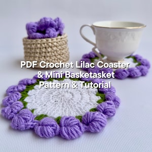 Puede incluir: Patrón de crochet PDF para un posavasos lila y una mini cesta. El posavasos es blanco con un borde de flores moradas y verdes. La cesta es beige con una flor morada en la parte superior.
