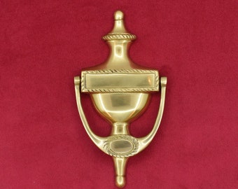 May include: A gold-toned door knocker with a rectangular nameplate and a decorative design. The knocker is shaped like a vase with a rounded top and a ring handle.