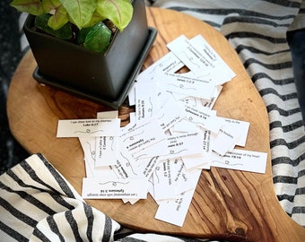 May include: A small potted plant with green and pink leaves sits on a wooden chopping board. Scattered around are small white cards with Bible verses and heart illustrations. A black and white striped cloth is in the background.