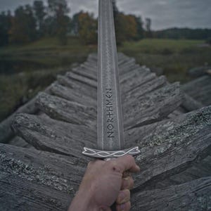 Może przedstawiać: Srebrny miecz z wygrawerowanym na ostrzu słowem "NORTHMEN". Miecz trzymany jest na tle drewnianego płotu i pochmurnego nieba. Rękojeść miecza ma dekoracyjny wz&oacute;r.