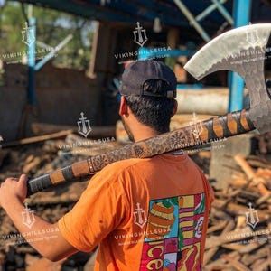 May include: A person wearing a bright orange t-shirt with a colorful graphic design is holding a large, hand-forged axe with a wooden handle. The axe has a hammered finish and a leather wrap on the handle. The axe is being held over the person's shoulder.