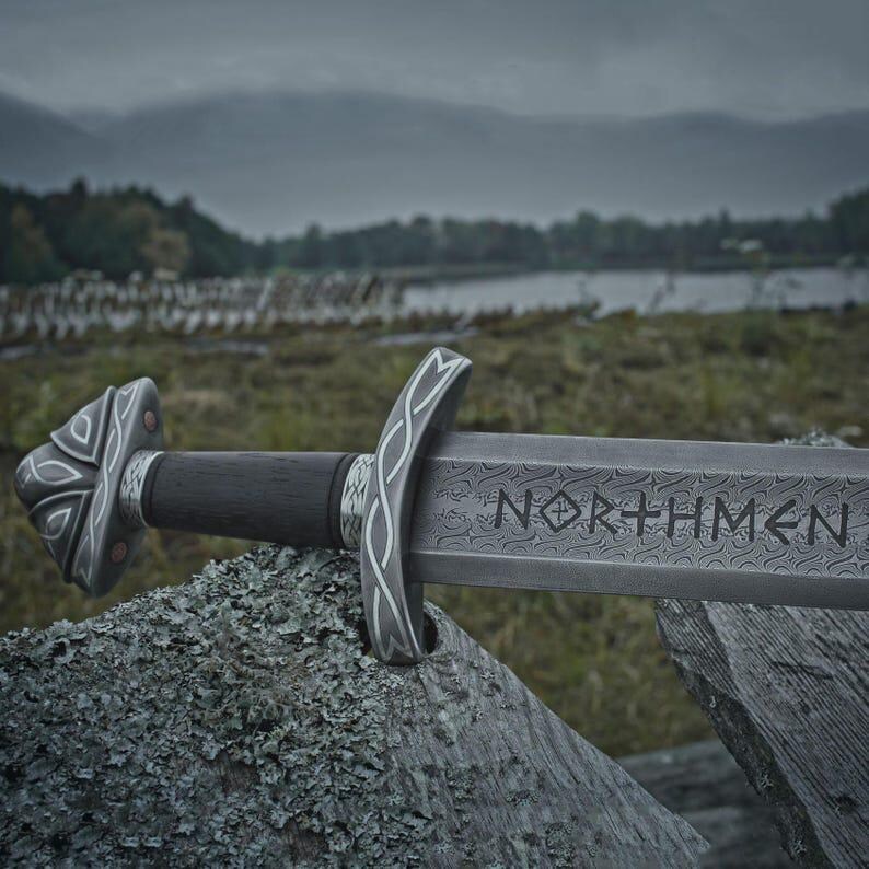 Może przedstawiać: Szczeg&oacute;łowe zbliżenie miecza z wygrawerowanym na ostrzu słowem "NORTHMEN". Miecz ma ciemny drewniany uchwyt, ozdobną srebrną rękojeść i osłonę oraz spoczywa na zwietrzałej drewnianej powierzchni. W tle widać jezioro i odległe wzg&oacute;rza.