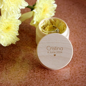 Könnte beinhalten: Eine hölzerne Ringbox mit einem Deckel, auf dem "Cristina 4. Juni 2024" und ein kleines Herz stehen. Die Box ist mit gelben Blütenblättern gefüllt und ein Ring befindet sich darin.