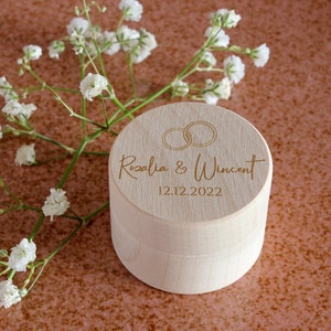 Könnte beinhalten: Eine Holzkiste für Ringe mit einem Deckel, der mit zwei ineinander verschlungenen Ringen, den Namen "Rosalia & Wincent" und dem Datum "12.12.2022" graviert ist.