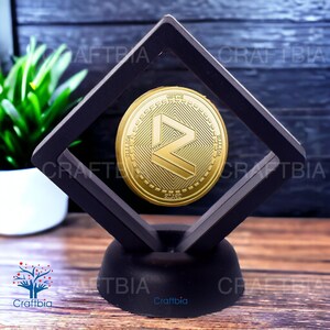 May include: A gold-coloured cryptocurrency coin with a black and white logo is displayed in a black diamond-shaped frame with a black base. The coin is suspended in the frame.