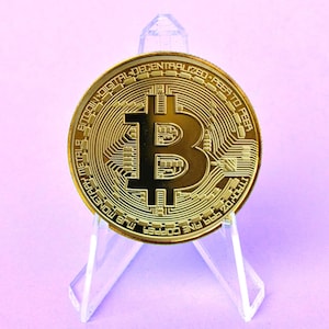 May include: A gold-coloured Bitcoin coin with the inscription "999 FINE COPPER MJB MONETARY METALS BITCOIN DIGITAL DECENTRALIZED P2P TO PEER 1 TROYOZ" displayed on a clear acrylic stand.