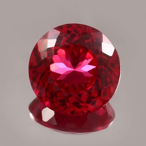 May include: A round, faceted gemstone with a deep, vibrant red color. The stone features a precise, symmetrical cut with multiple triangular and diamond-shaped facets that create internal light reflections and a brilliant, sparkling appearance.