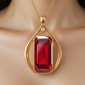 May include: A gold pendant necklace featuring a large, rectangular, deep red gemstone set within a smooth, teardrop-shaped gold frame. A small, clear, round-cut stone is positioned at the top of the frame, connecting to a delicate gold chain.