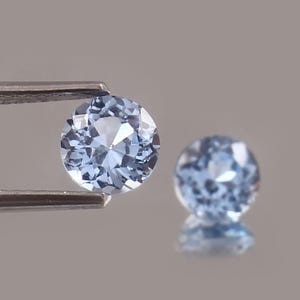 May include: Two round, light blue gemstones. One is held by a metal tweezer, and the other is slightly out of focus in the background. The gems have a faceted cut, reflecting light. The background is a neutral gray.