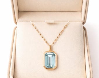 May include: A gold necklace with a rectangular, light blue gemstone pendant, displayed inside a beige jewelry box. The pendant is set in a gold frame and hangs from a delicate gold chain. The box is open, revealing a soft, cream-colored interior.