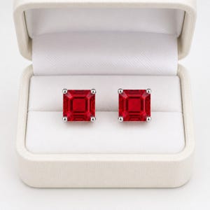 May include: A pair of square-cut red gemstone stud earrings with silver-toned metal prongs, presented inside a cream-colored jewelry box with a white interior lining. The earrings feature a faceted design that reflects light across the surface.