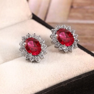 May include: A pair of stud earrings featuring a central oval-cut vibrant red gemstone surrounded by a halo of small, clear, round-cut white stones set in a silver-toned metal frame, presented in a cushioned jewelry box with a black exterior.