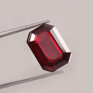 May include: A deep red, emerald-cut gemstone held by silver-toned metal tweezers. The stone features a rectangular shape with faceted edges that reflect light, creating a clear, transparent appearance with dark and bright crimson color gradients.