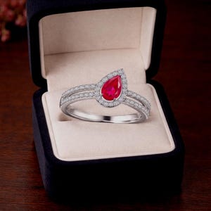 May include: A silver ring with a pear-shaped, vibrant red gemstone, surrounded by a halo of smaller, clear stones. The ring features a split band, and is presented in a black jewelry box with a cream-colored interior.