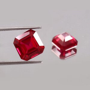 May include: Two vibrant red, square-cut gemstones with faceted surfaces. One gemstone is held by silver-toned metal tweezers, while the other rests nearby, displaying a clear reflection on the smooth surface beneath it.
