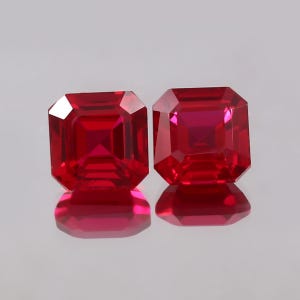 May include: Two radiant-cut, deep red gemstones. Each gem has a square shape with beveled edges, reflecting light. The gems are displayed on a reflective surface, creating mirror images. The color is a rich, saturated red.