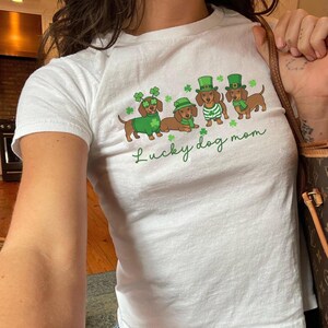 Pode incluir: Camiseta branca com tema do Dia de São Patrício. Apresenta quatro dachshunds usando chapéus e roupas verdes, com o texto "Lucky dog mom" em escrita verde. Os cães são marrons e verdes.