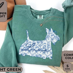 Pu&ograve; includere: Felpa verde chiaro con un motivo floreale a forma di cane e il nome "Luna". La felpa ha un girocollo e maniche lunghe. L'immagine include anche il testo "Super Soft", "Comfort Colors", "Light Green", "5 Sizes" e "8 Colors".